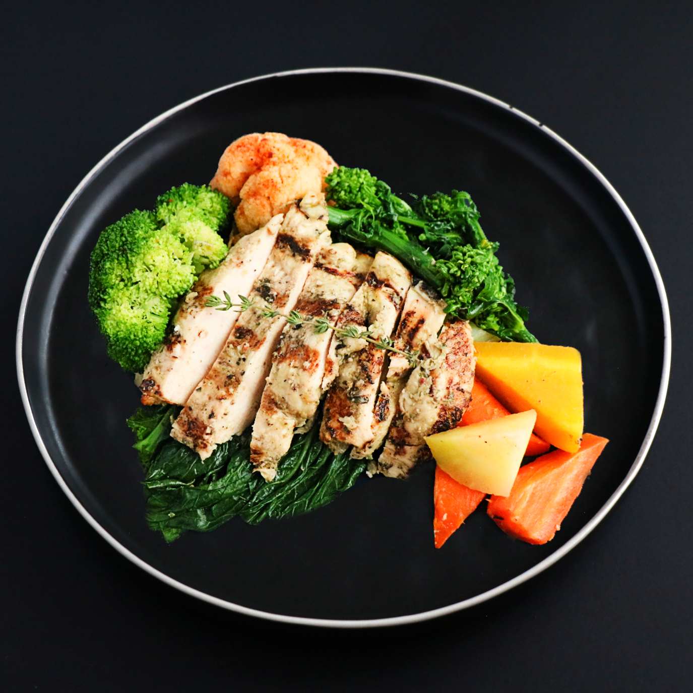 Sliced grilled chicken breast with assorted vegetables around on a black plate