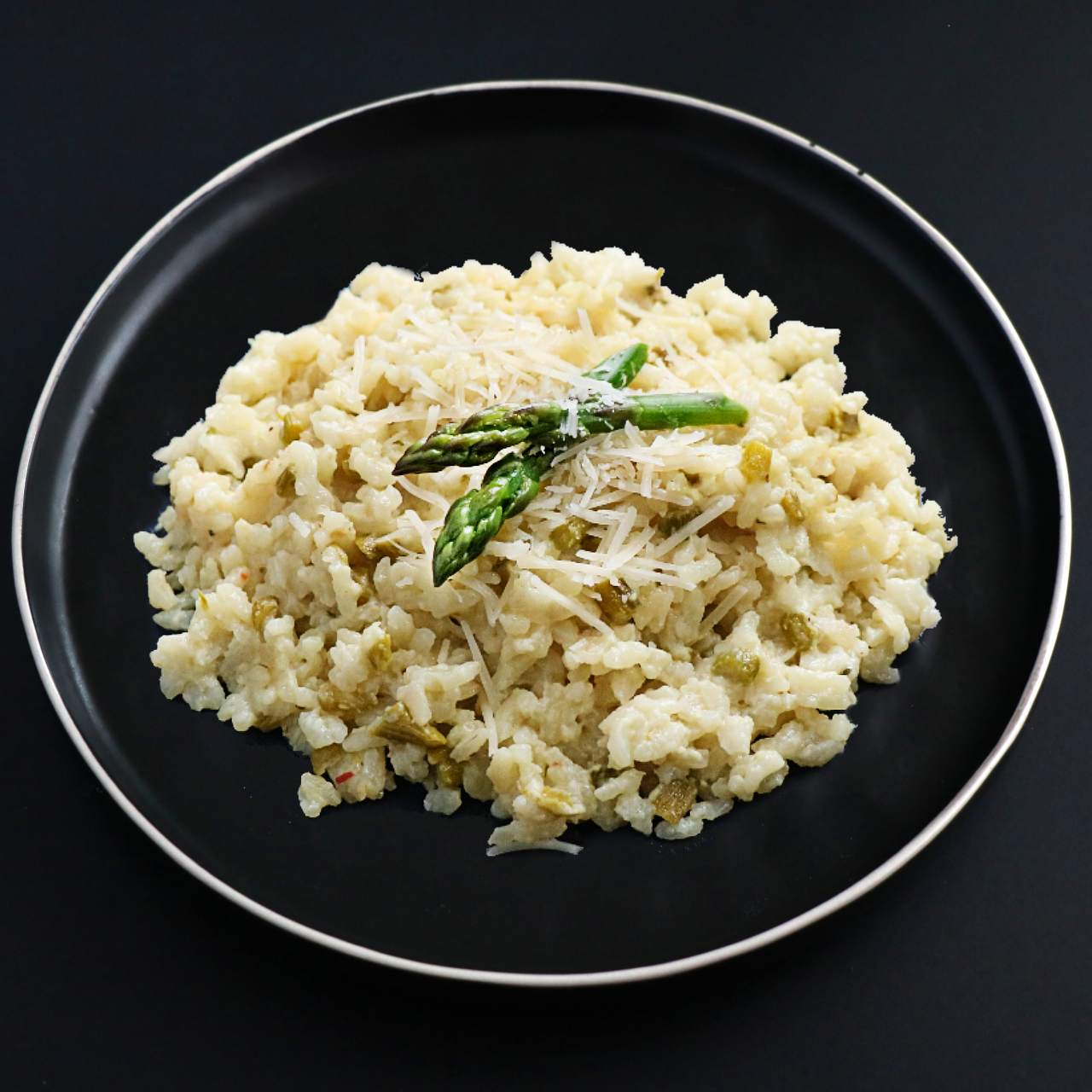 Creamy and luscious asparagus risotto topped with asparagus pieces and vegan cheese on a black plate.