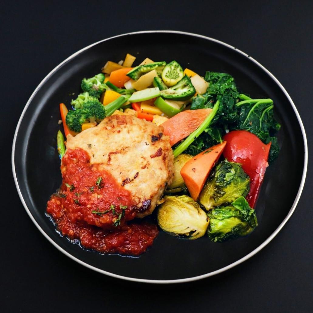 Mediterranean Turkey slider from balanced menu is slightly covered with tomato jam and herbs placed on a black plate surrounded by assortment of vegetables
