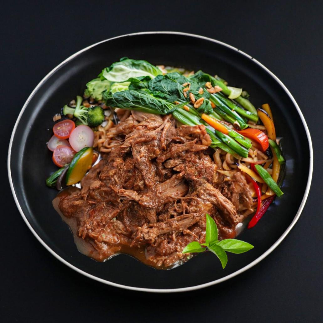 A plate of Red Thai Pulled Beef Curry with a side of assorted oriental vegetables, presented on a dark plate garnished with a leaf of thai basil.