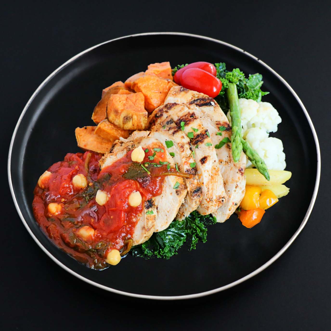 A plate of gourmet Tuscany Chicken meal with roasted sweet potato, asparagus, and tomato, presented on a black round dish.