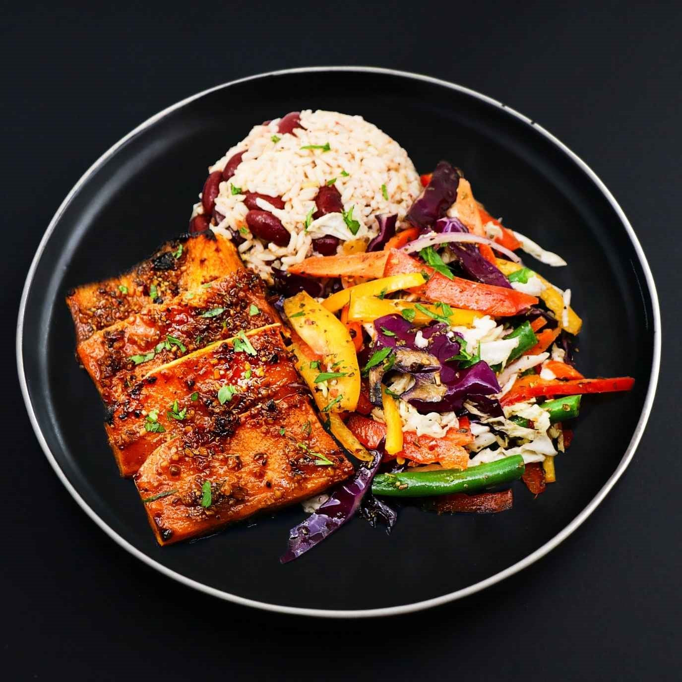 Pieces of plant-based chicken alongside black bean brown rice and assortment of vegetables on a black plate.