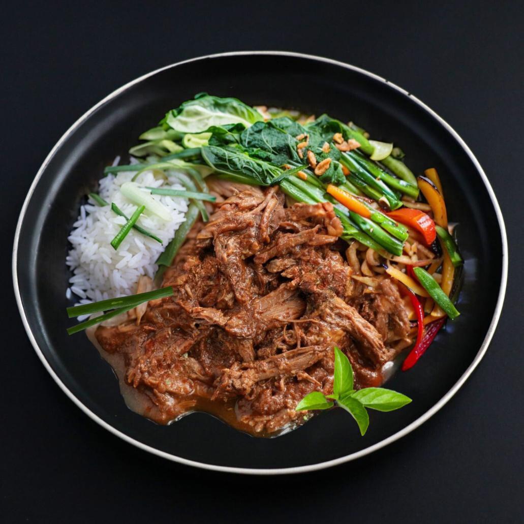A dish of aromatic and tender pulled beef covered with lucious red Thai curry sauce, sided with jasmine rice and variety of veggies with a Thai basil leaf for garnish represents the gourmet menu.