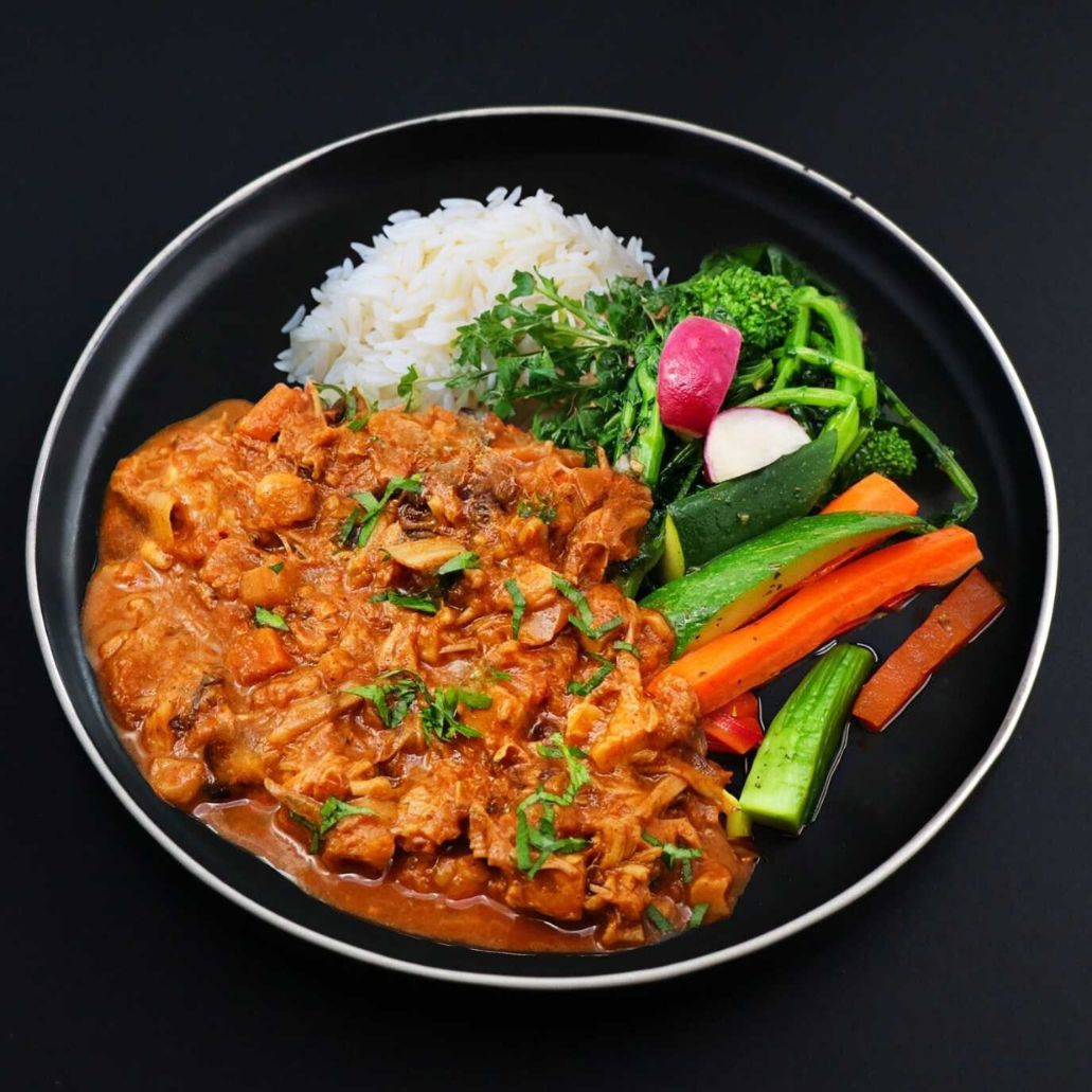 Red Thai jackfruit curry dish garnished with parsley and complemented with a side of jasmine rice and assortment of vegetables on a black plate.