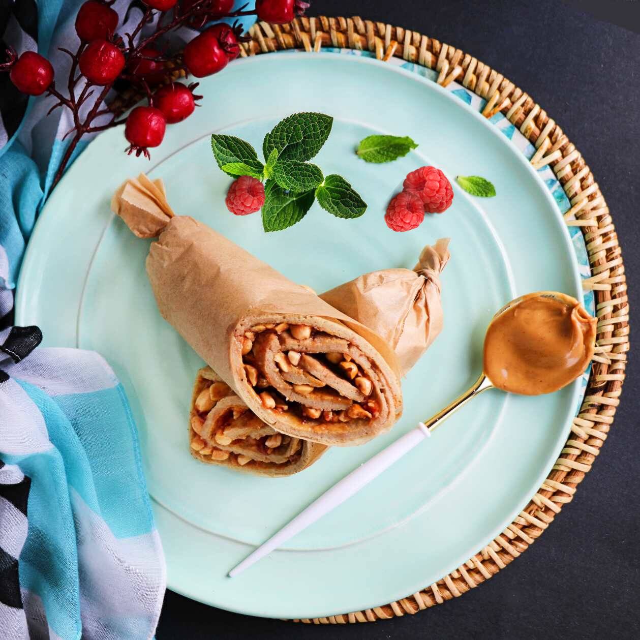 A vegan oatmeal crepe roll filled with creamy peanut butter and raspberry jam, garnished with mint leaves and a drizzle of maple syrup on a blue plate with a side of berries and a golden spoon.