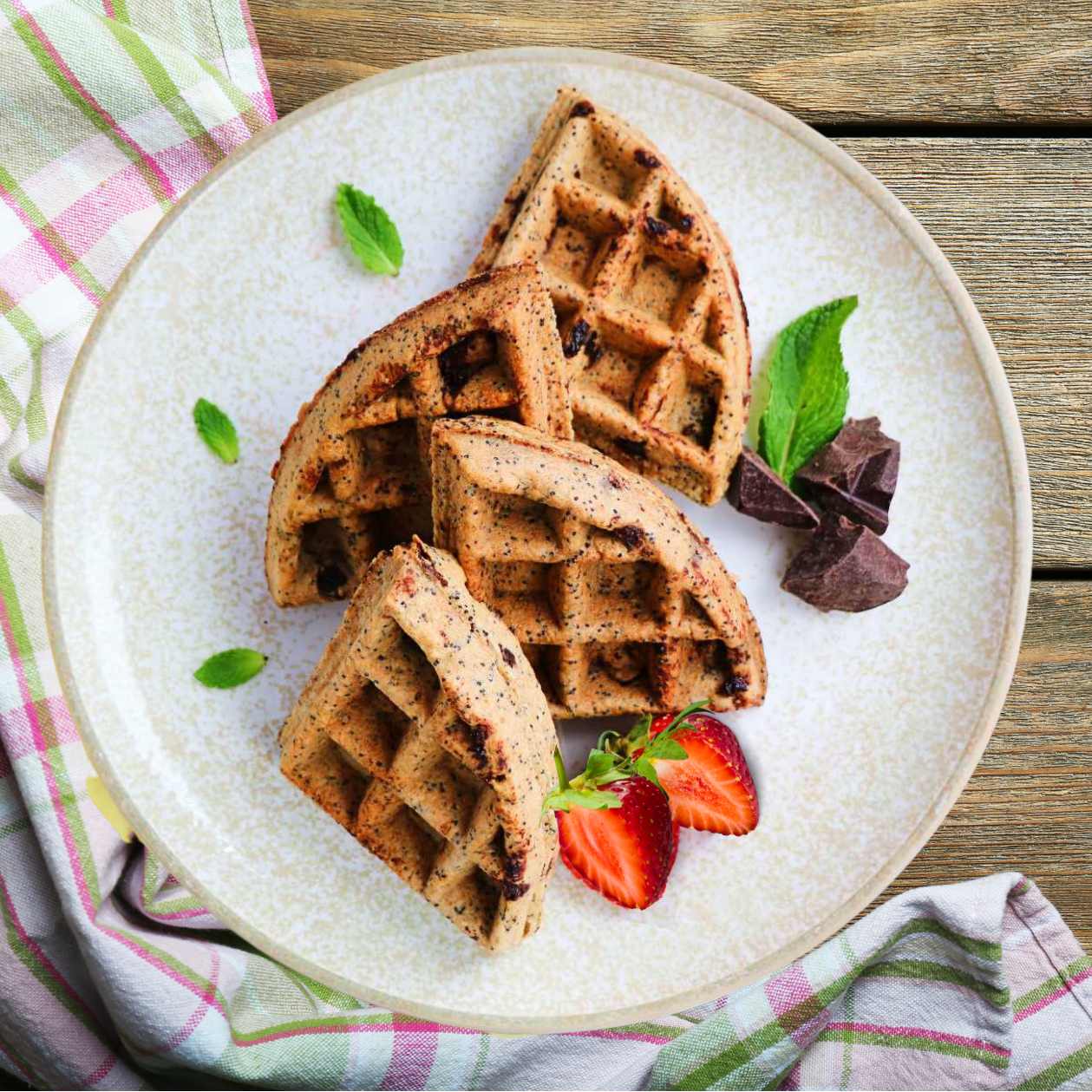 A plate with three chocolate chip vegan waffles, garnished with poppyseeds, a slice of strawberry and a mint leaf on the side, on a wooden table with a striped pink and green napkin to the side.