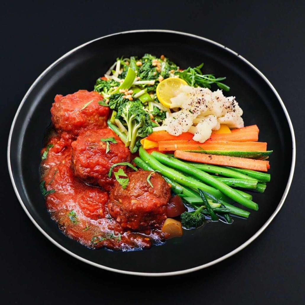 A plate with juicy Italian meatballs in a tomato sauce, accompanied by steamed green beans, sliced carrots and other veggies.