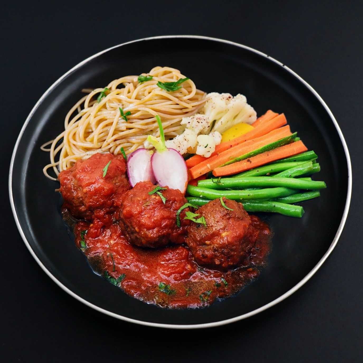 A plate of Italian meatballs in tomato sauce, served with spaghetti and a variety of vegetables including green beans, carrots, and onions represents one of the gourmet menu dishes.