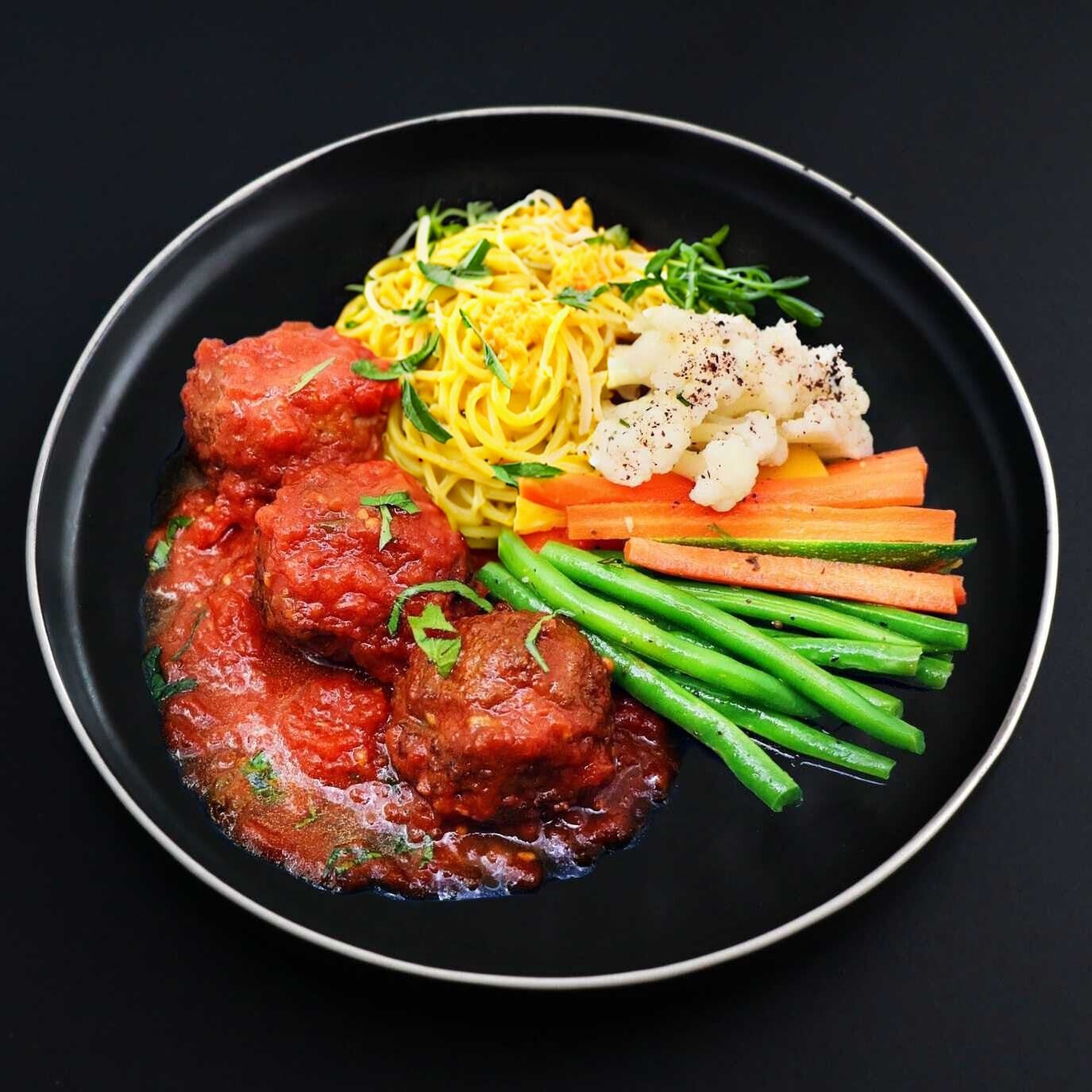 Juicy Paleo Italian meatballs under lucious tomato sauce sided with spaghetti squash noodles and assorted veggies on a black plate.