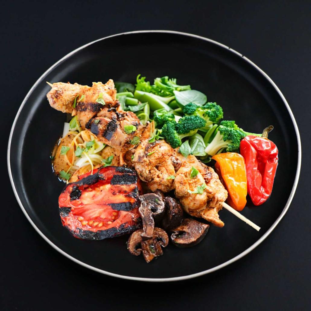 A chicken shish kebab skewered with a piece of broccoli, another of tomato, mushroom, and a slice of red bell pepper on a black plate, garnished with parsley.