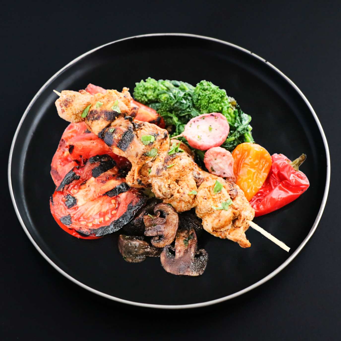 A chicken shish kebab skewered with onion and pepper, grilled and served on a black plate with a side of a grilled tomato