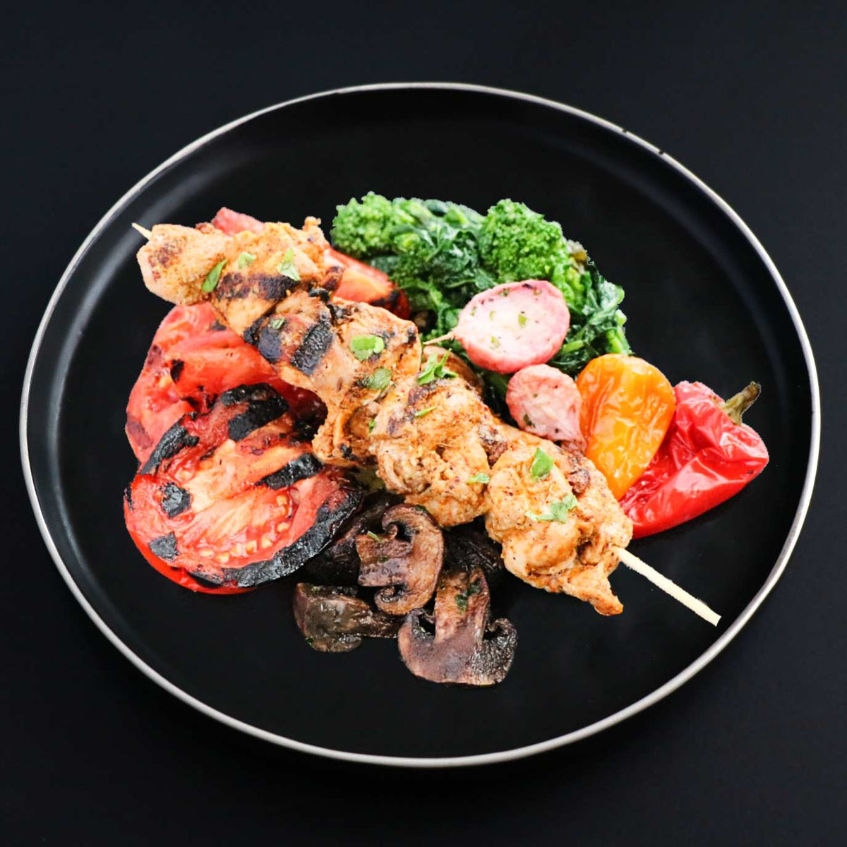 A chicken shish kebab skewered with onion and pepper, grilled and served on a black plate with a side of a grilled tomato