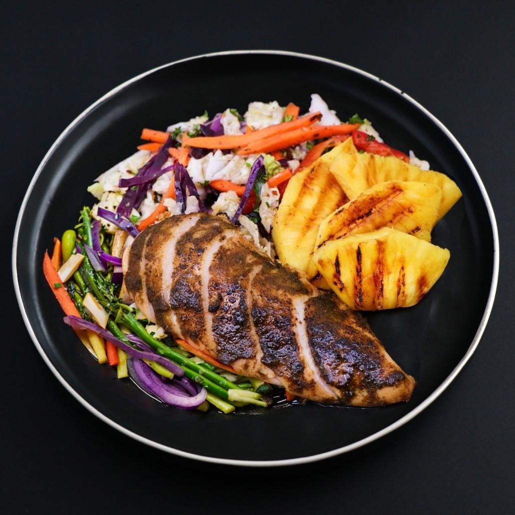 Paleo Jerk chicken breast is sliced and placed next to grilled pineapple pieces and assorted vegetables. 