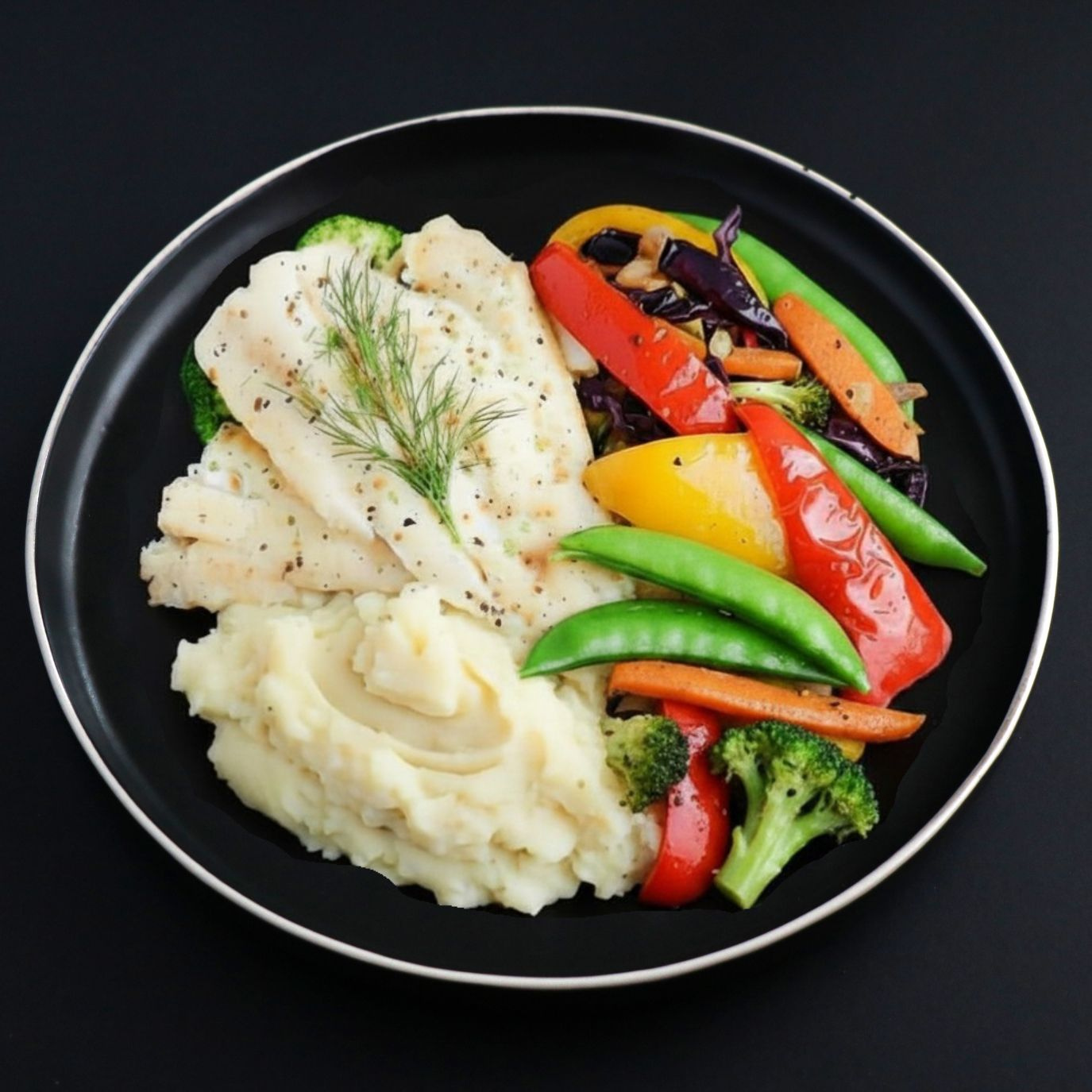 A plated meal featuring cooked white fish fillets, mashed potatoes with chopped chives, and mixed vegetables including broccoli, cauliflower, carrots, and red and yellow bell peppers on a black plate against a dark background.