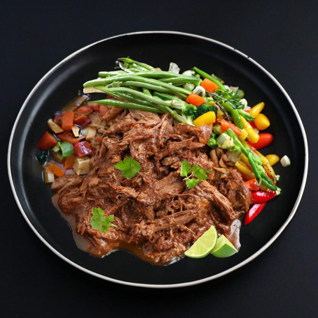 A plate of beef barbacoa with assorted vegetables and lime wedge on the side.