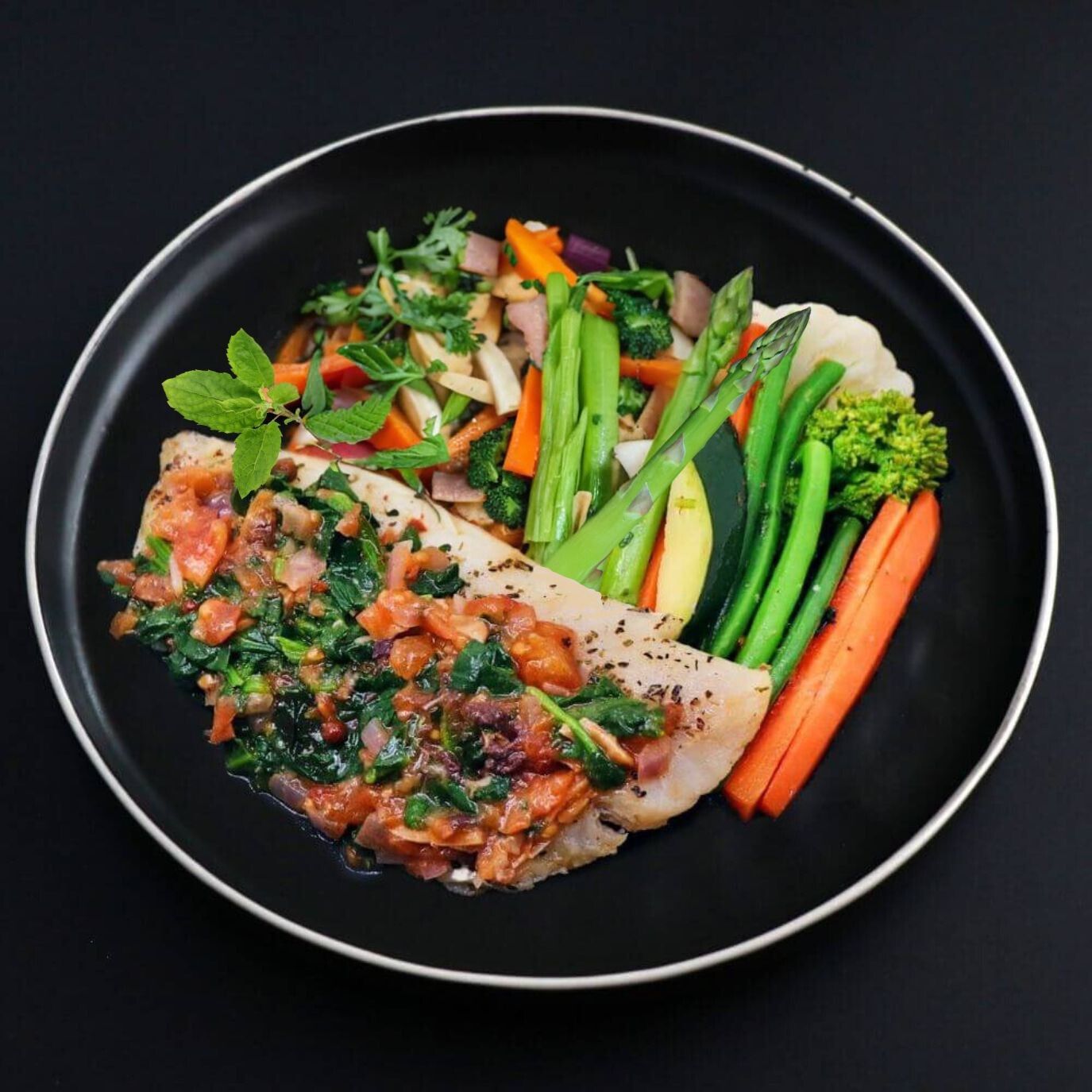 A piece of a tender Cod fillet is nestling on a black plate complemented with a variety of seasonal vegetables and covered with aromatic tomato-spinach Spanish sauce.