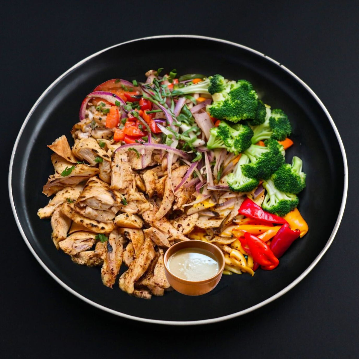 Balanced turkey shawarma bowl dish presented on a black plate along with a side of assorted vegetables, herbs and homemade tahini