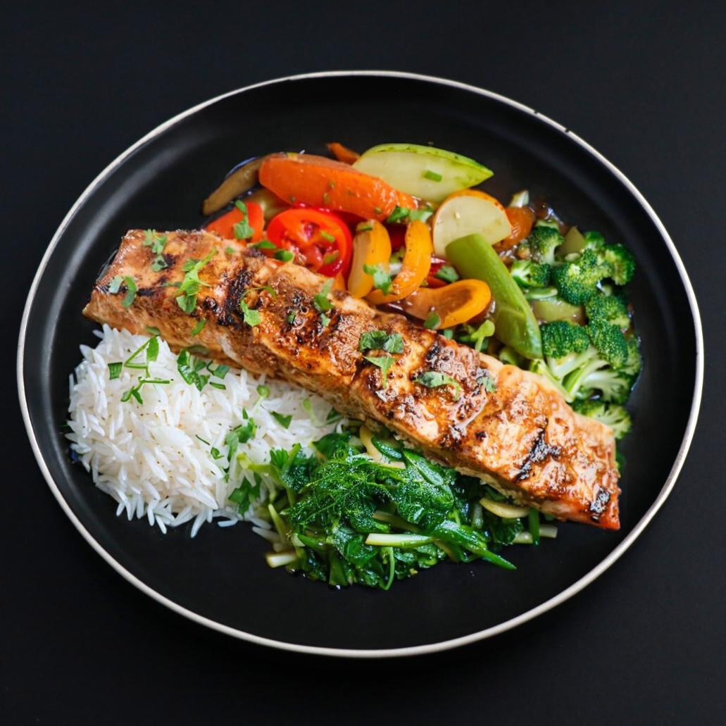 Juicy slice of gourmet tandoori salmon atop basmati rice complemented with assorted veggies and leafy greens.