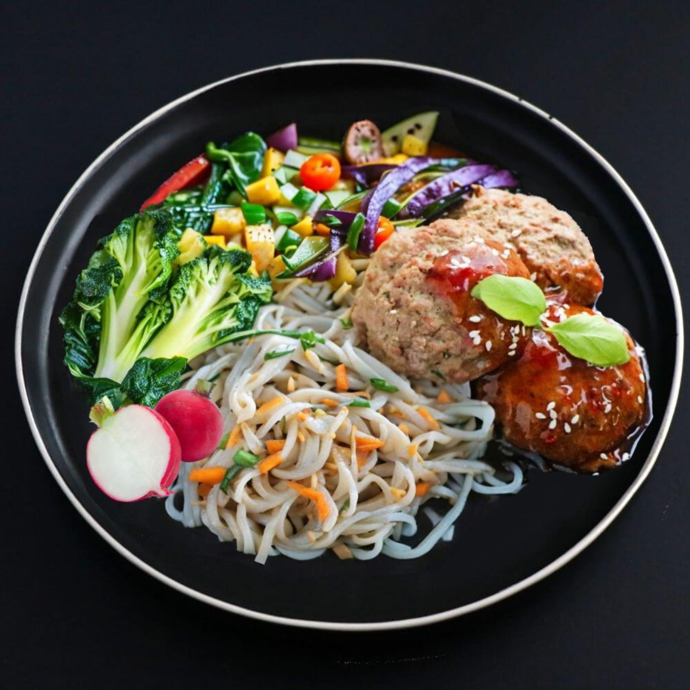 A plate containing Asian turkey meatballs, sweet potato noodles, and sautéed greens, presented on a dark background.