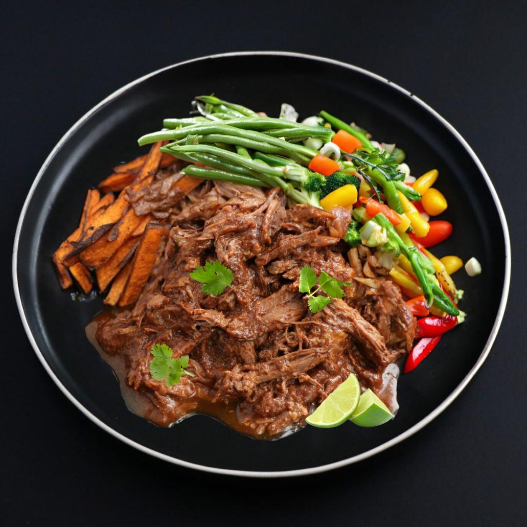 A plate of beef barbacoa with sweet potato fries, garnished with green onions, side lime wedge and cilantro, served on a dark plate against a black background.