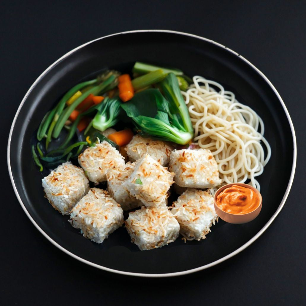 A black plate containing organic crispy coconut crusted tofu, sweet potato noodles, and seasonal vegetables, drizzled with sriracha vegan mayo.