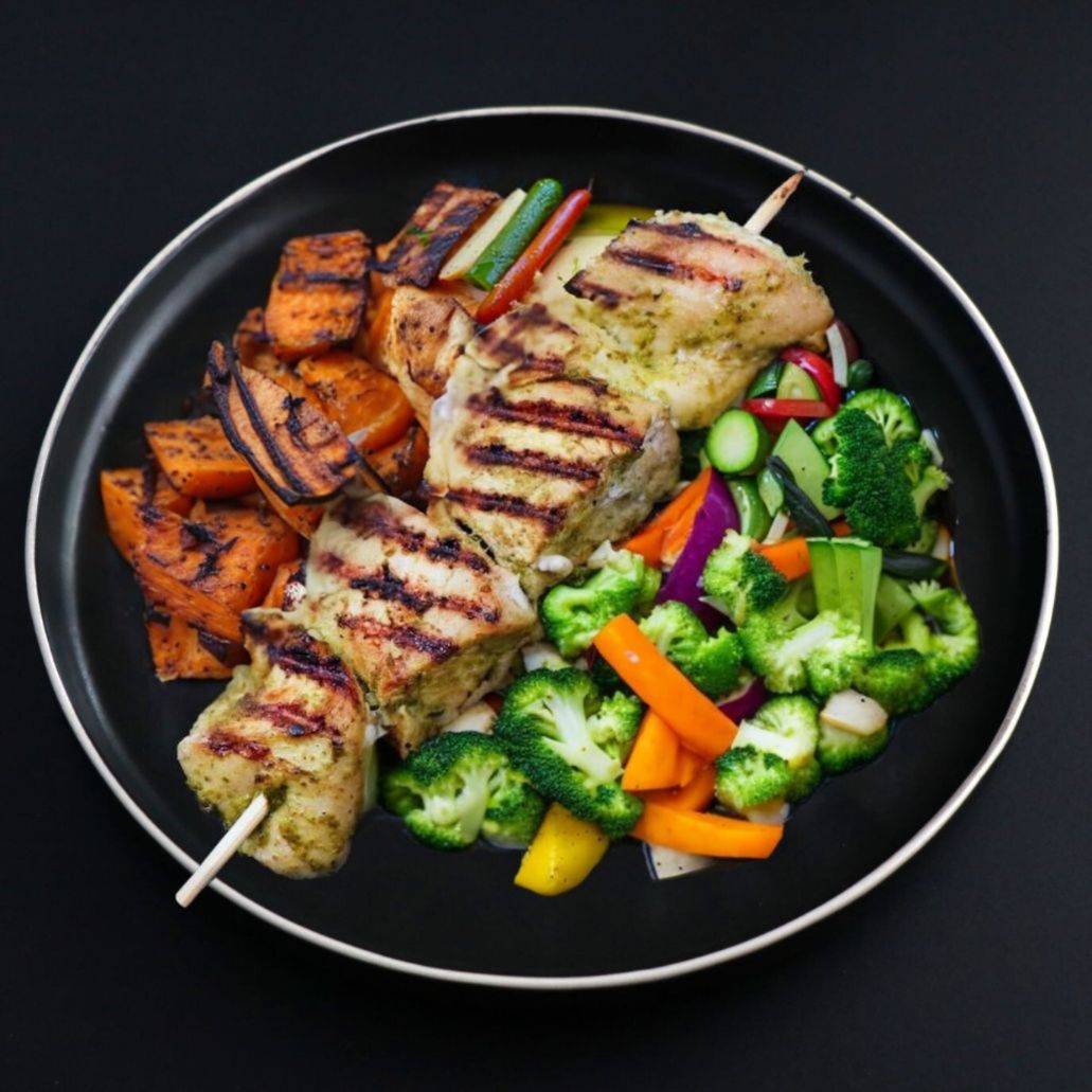 A plate containing grilled chicken souvlaki skewers with roasted sweet potatoes and a side of broccoli and carrots and other veggies.