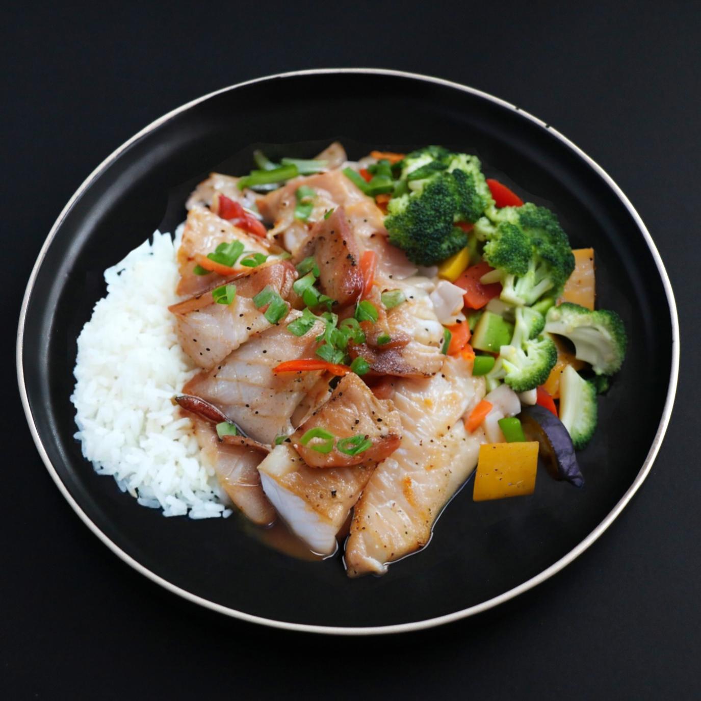 A prepared meal consisting of miso glazed cod, jasmine rice, and a side of assorted vegetables on a black plate.