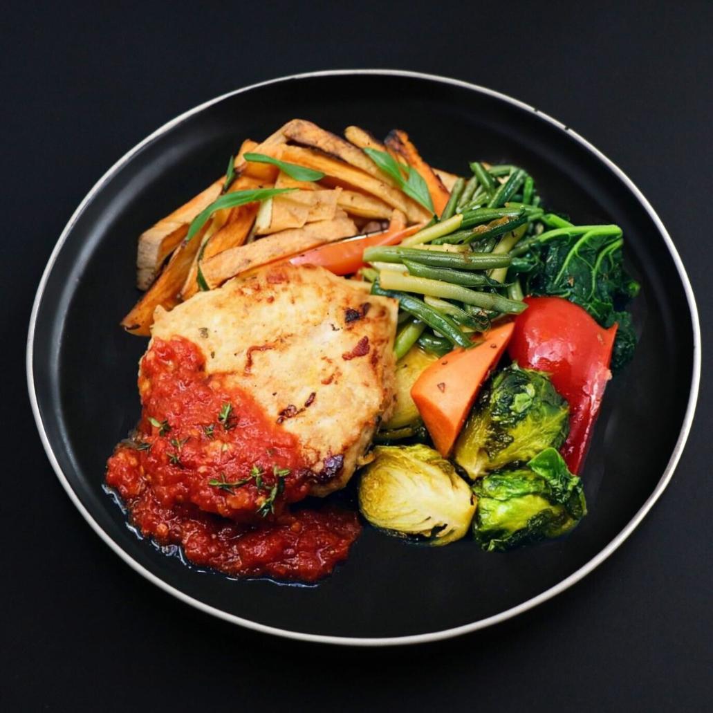 Black plate with paleo Mediterranean turkey slider slightly covered with homemade tomato jam and herbs, paired with roasted potato fries and assortment of vegetables. 