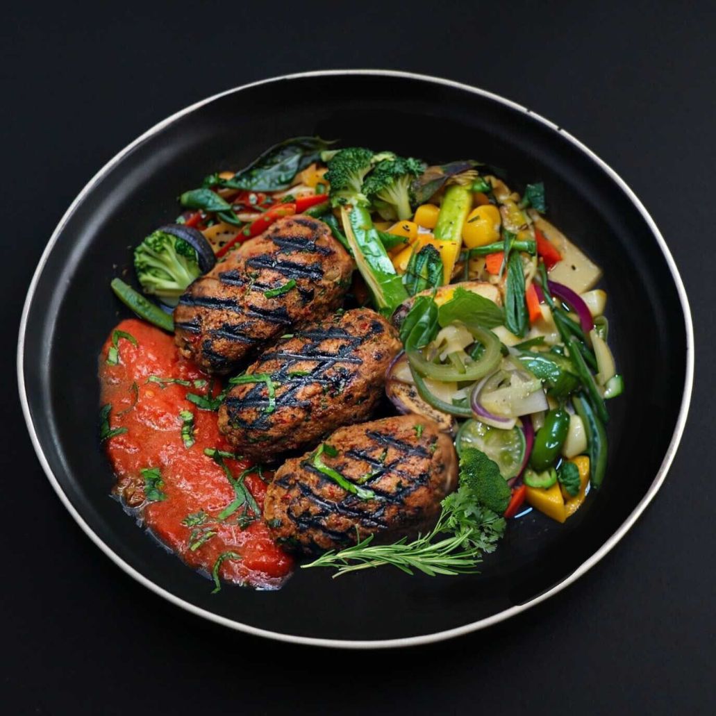 Dark plate featuring aromatic balanced beef kafta kebabs sided with seasonal veggies, herbs and homemade tomato sauce
