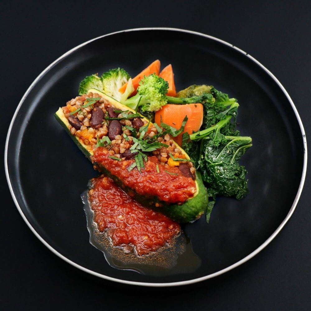 Vegan beef stuffed zucchini dish on a black plate, complemented with a side of assorted vegetables and topped with a luscious homemade tomato sauce. 